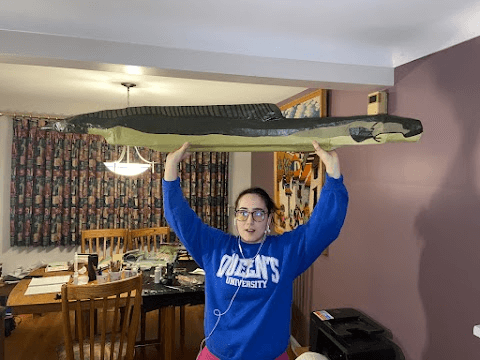 Claudia, sitting next to a cluttered dining room table, holds the eel sculpture aloft in both hands. 