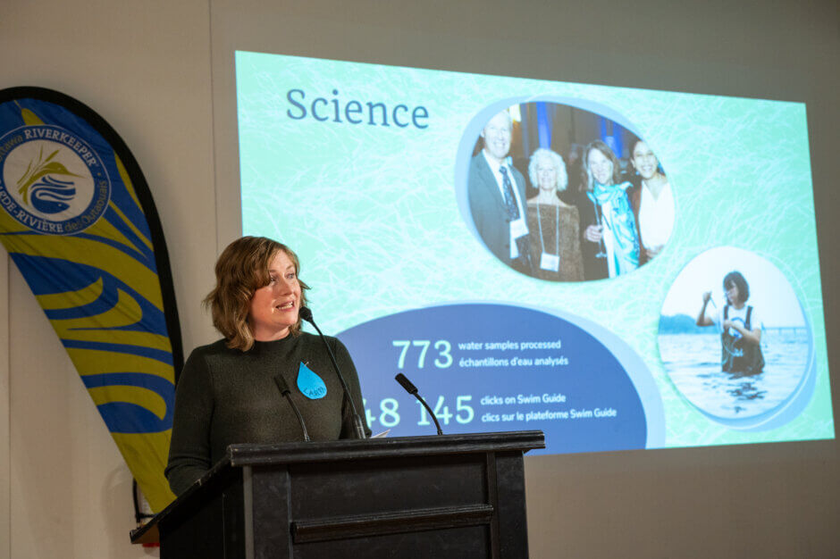 Laura Reinsborough, Riverkeeper and CEO, speaks to the audience at the 2023 Annual Public Meeting. The slide behind her is titled "Science" and shows that Ottawa Riverkeeper collected 773 water quality samples this year, with photos showing staff collecting samples. 