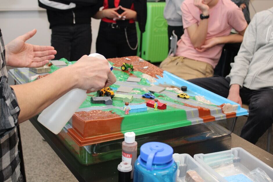 A closeup of the watershed model, which shows it covered with various toy buildings and cars. There are students watching in the background as someone in the foreground explains and holds a spray bottle to simulate rain. 