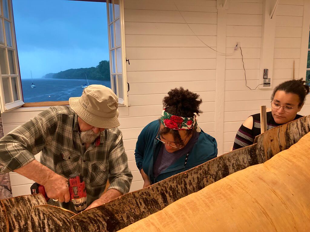 Pinock Smith works on a large section of birch back as two youth participants assist. The background shows a view out the window of the Ottawa River. 