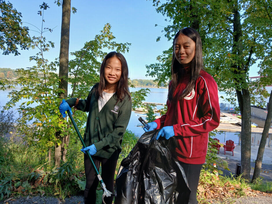 Two young students collecting garbage as part of a cleanup, wearing gloves and holding a trash bag and garbage picker. 