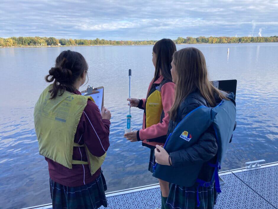 Three students wearing lifejackets standing on the deck of River house. One is testing the water with a sampling stick and conductivity meter, while another holds a clipboard of data. 