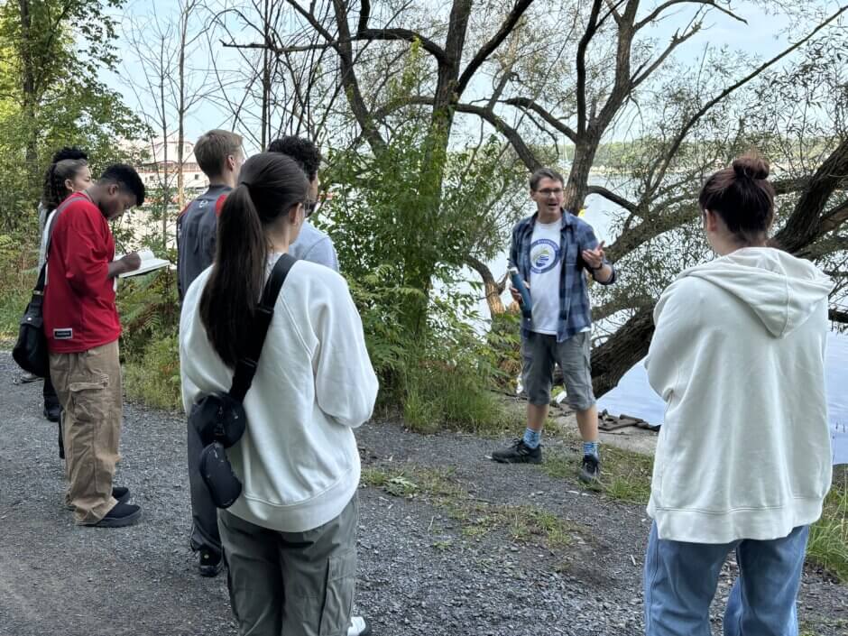 Youth standing outside along the banks of the river listening to a presentation. 