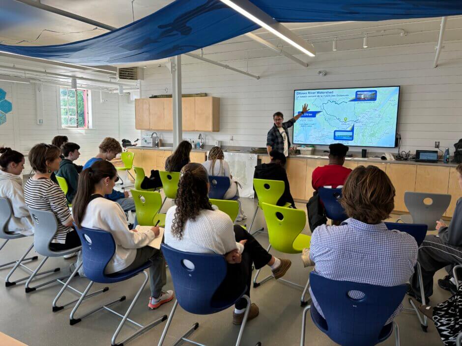 A River School workshop, showing students in rows of chairs looking at an educator pointing to a map of the Ottawa River watershed on a large screen in the Ottawa Riverkeeper Learning Lab. 